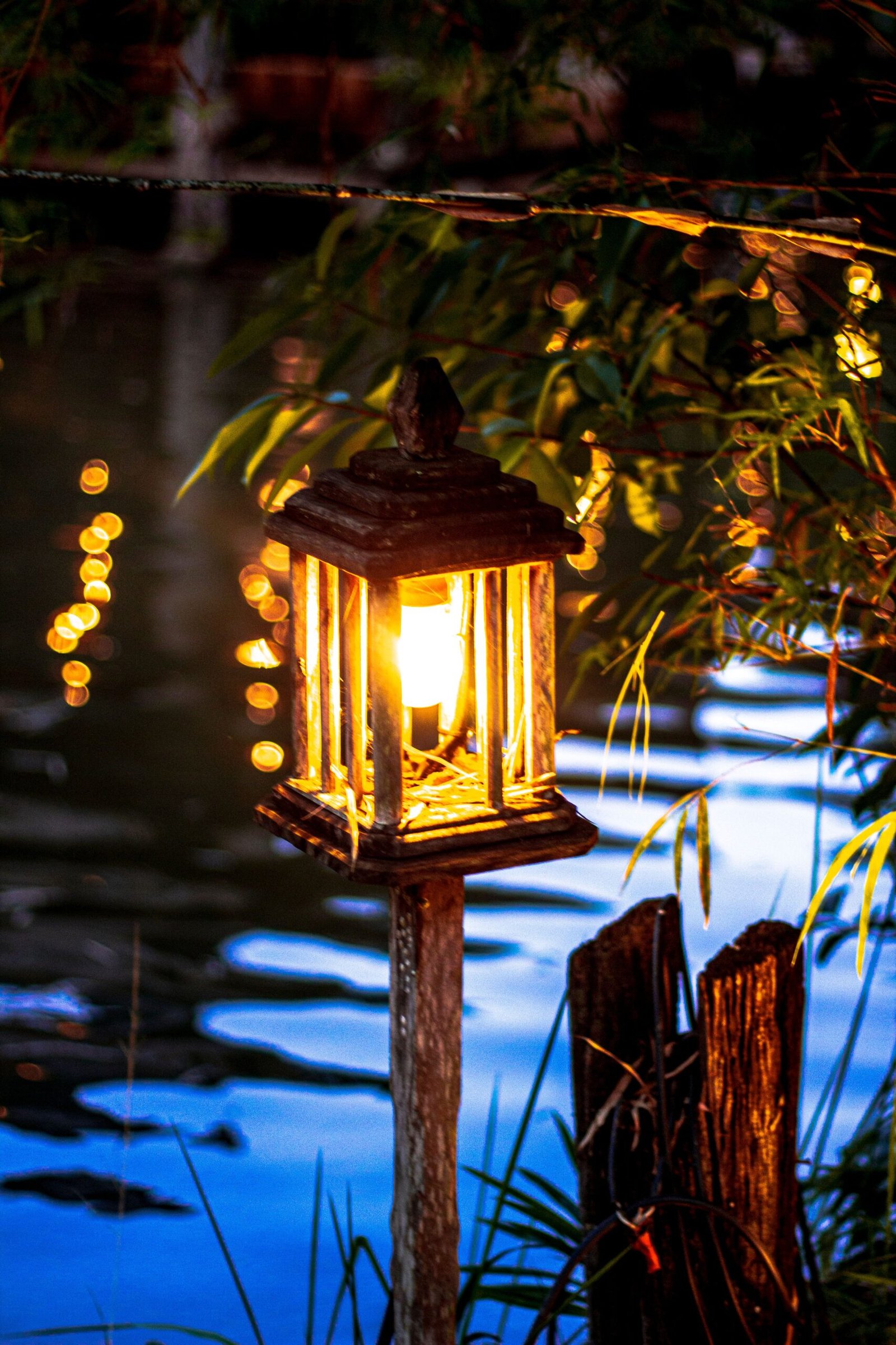 A lantern glows warmly by a tranquil lakeside, creating a peaceful and magical atmosphere at dusk through twinkling lights.