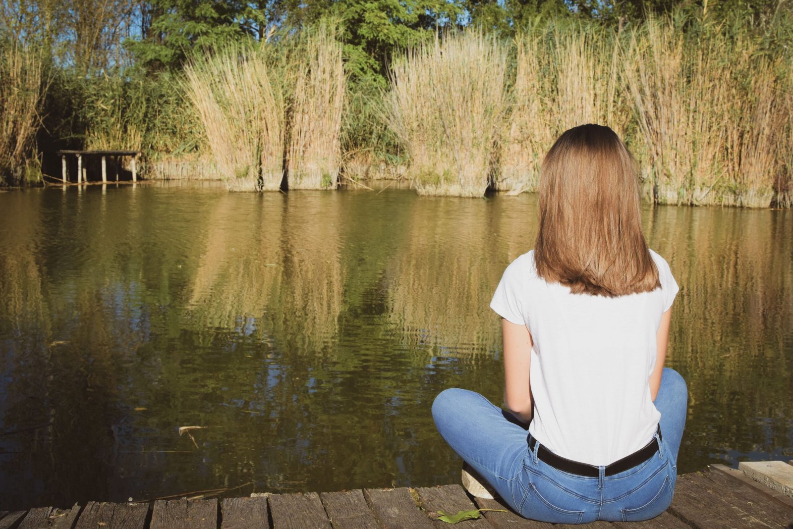 A young woman sits on a wooden pier by the lake, surrounded by tall grass.